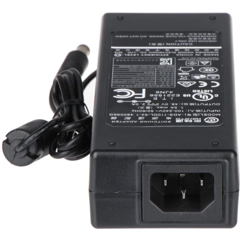 Na zdjęciu znajduje się czarny zasilacz sieciowy z widocznymi oznaczeniami technicznymi i certyfikatami na obudowie. Zasilacz posiada kabel z wtyczką do podłączenia do urządzenia elektrycznego.