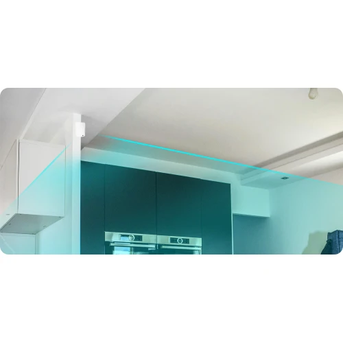 Zdjęcie przedstawia wnętrze nowoczesnej kuchni z zamontowaną w rogu bezprzewodową czujką ruchu. Widać także niebieską linię ilustrującą zasięg działania czujki.