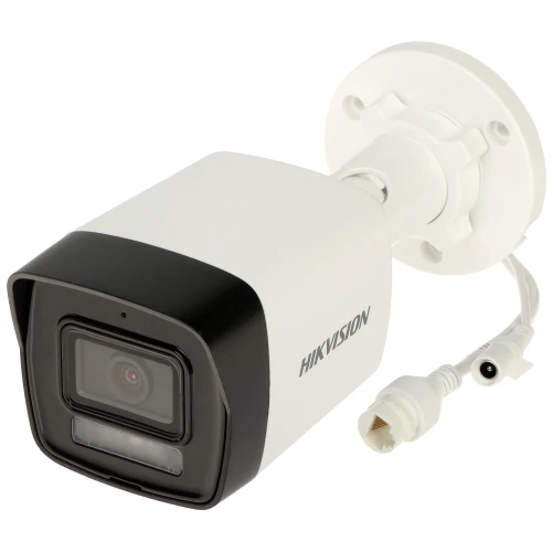 Na zdjęciu znajduje się biała kamera monitoringu marki Hikvision z czarnym frontem, zamontowana na białym uchwycie ściennym. Obok kamery widoczne są dwa przewody z wtyczkami do podłączenia.